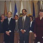 Janet Reno and Shay Bilchik in an official setting with flags behind them.