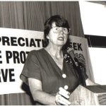 Janet Reno giving a speech at a podium with a poster behind her.