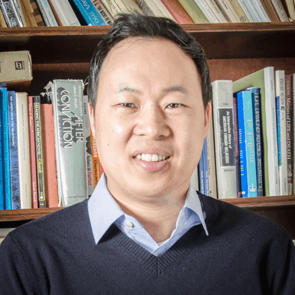 Lewis Lee smiling in sweater with bookcase behind him