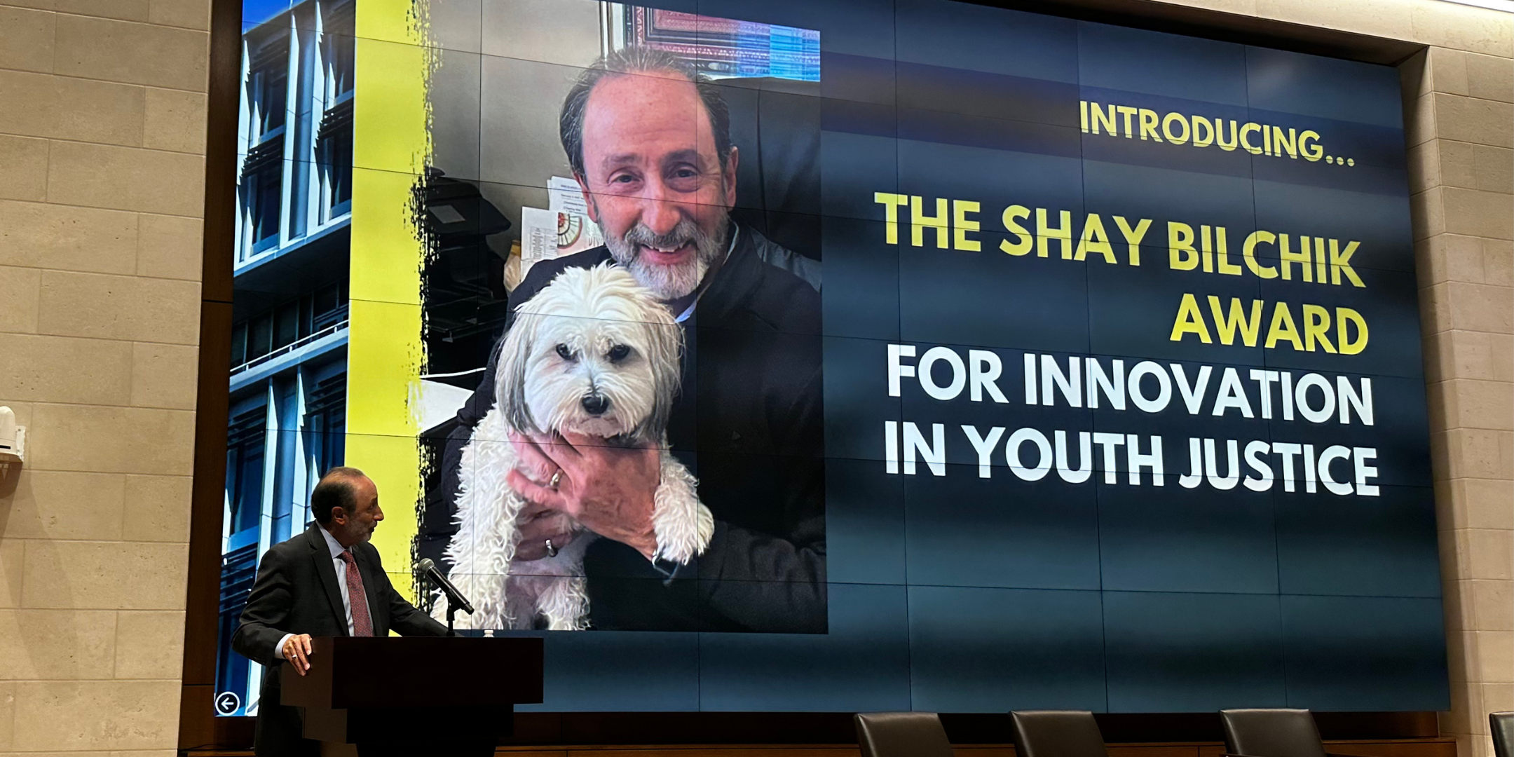 Shay at podium speaking in front of large screen